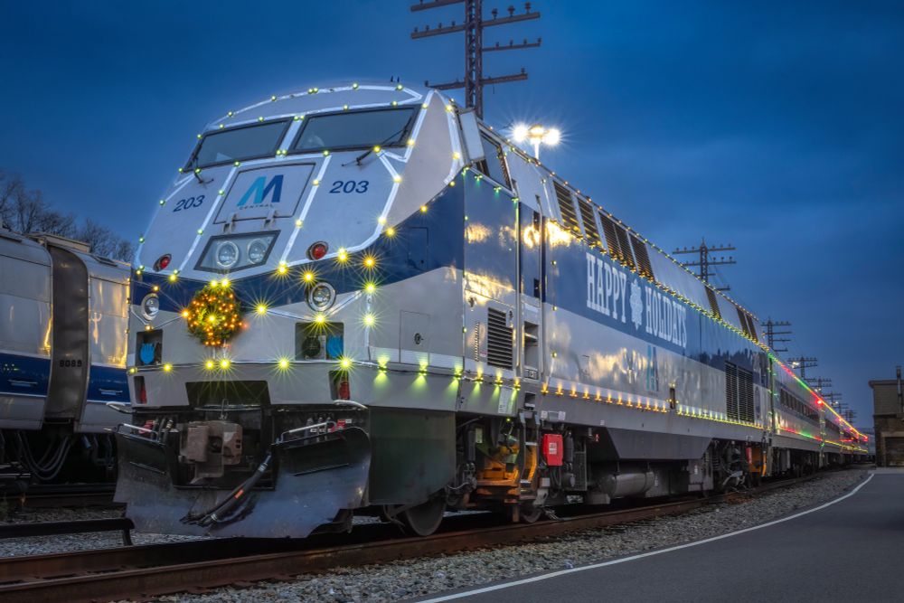 The Holiday Lights diesel train, decorated with green and red string lights and a wreath in front of the train, stationed by the maintenance shop in the early evening.