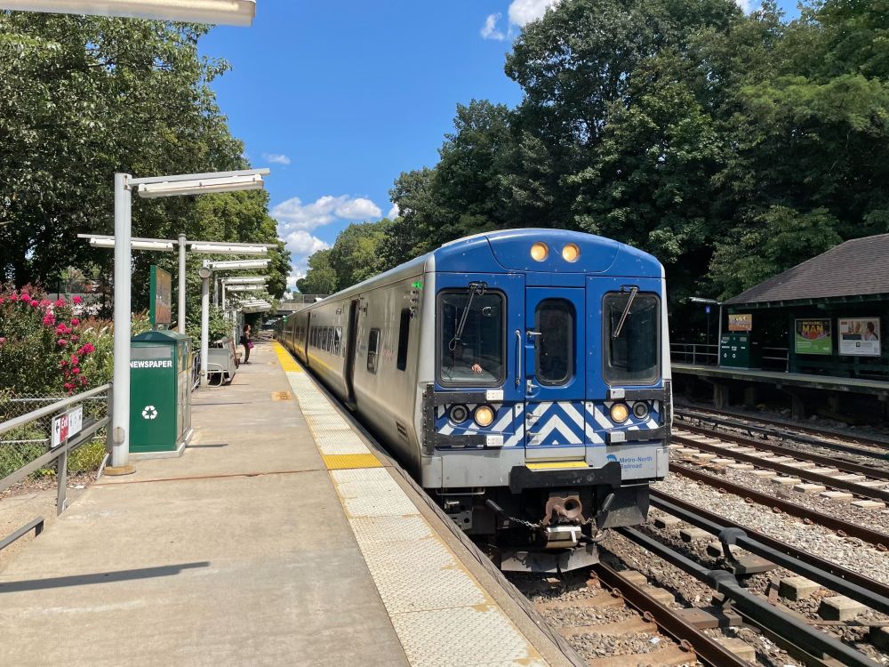 A Metro-North train pulling into Botanical Garden station on a sunny day, with a person waiting on the platform.