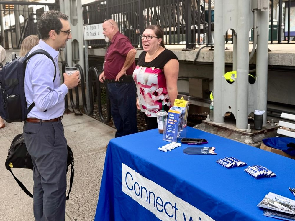 A Metro-North employee speaking with a customer at the Connect with Us table, lined with pens and brochures. 