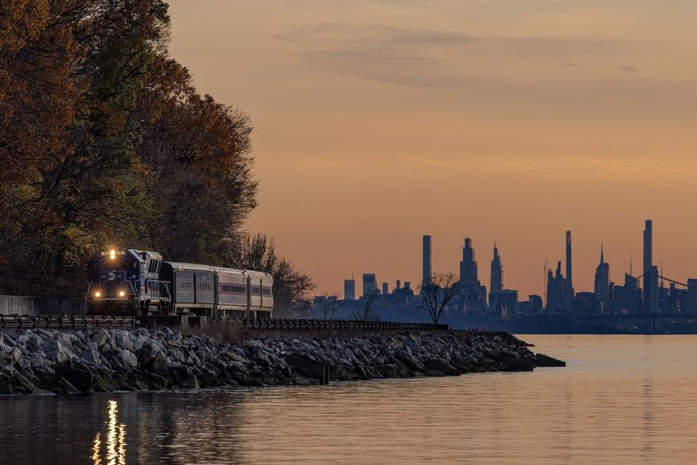 A distant shot of a Metro-North train running along the tracks next to the Hudson River, with the cityscape in the background at sunset.
