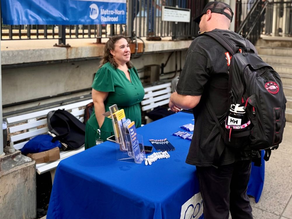 A Metro-North employee speaking with a customer at the Connect with Us table, lined with pens and brochures. 