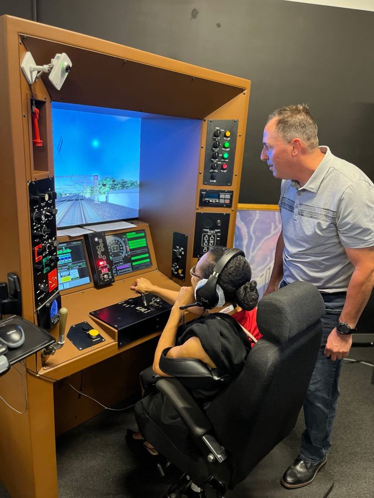 A Metro-North instructor guiding a COMTO attendee wearing headphones and navigating a virtual reality demo on a large screen. 