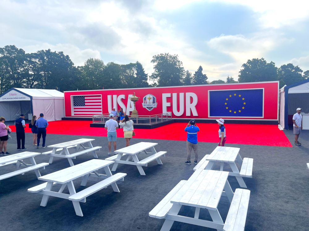 People taking pictures of a large red display that reads "USA" and "EUR" with their respective flags, on a cloudy day. There are several white benches behind them.