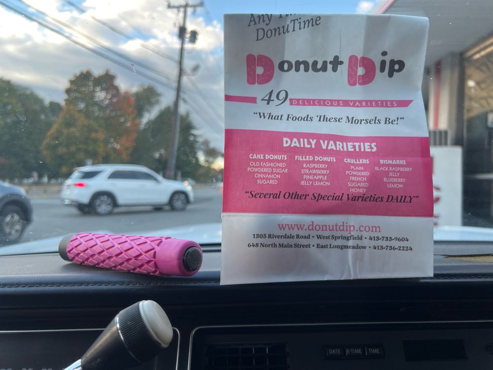a collapsing baton with a pink handle and a bag from Donut Dip (containing a bismarck) sit on the dashboard of a very old car