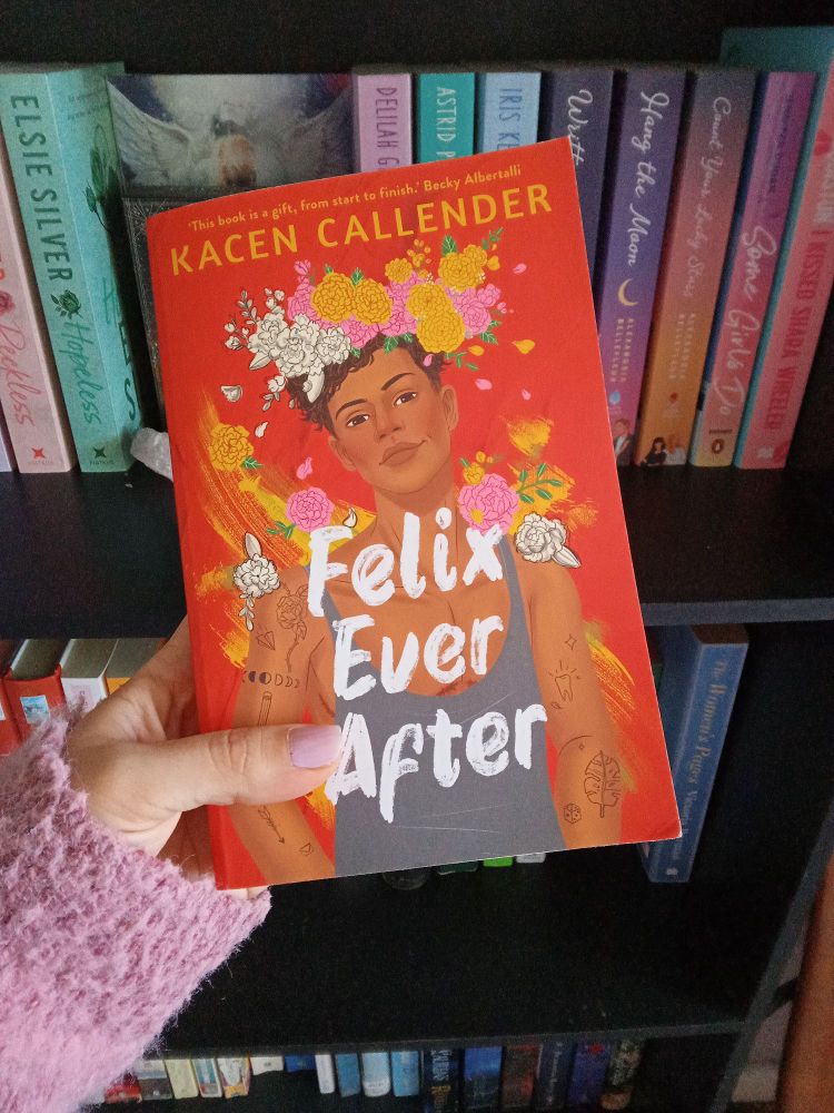 Picture of a hand holding a book, Felix Ever After by Kacen Callender, with a bookshelf in the background.