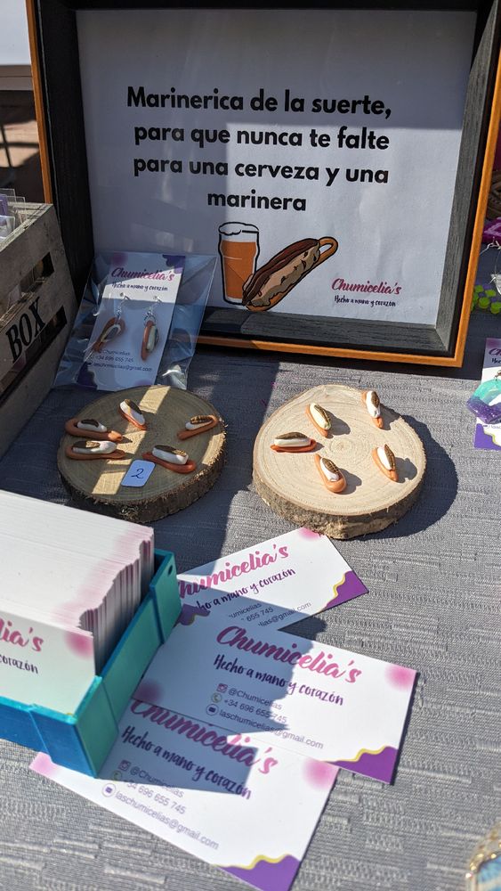 Stand de mercadillo en el que se ven dos soprtes con unas marineras pequeñas hechas de arcilla polimérica. Hay un pequeño cartel que se lee: "Marinericas de la suerte, para que nunca te falte para una marinera y una cerveza"