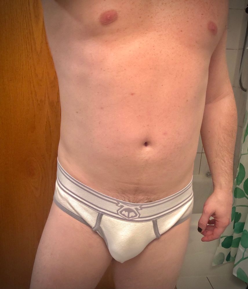 A man standing in white underwear with a prominent bulge and visible penis line. Upper legs, belly, and chest are visible. 
