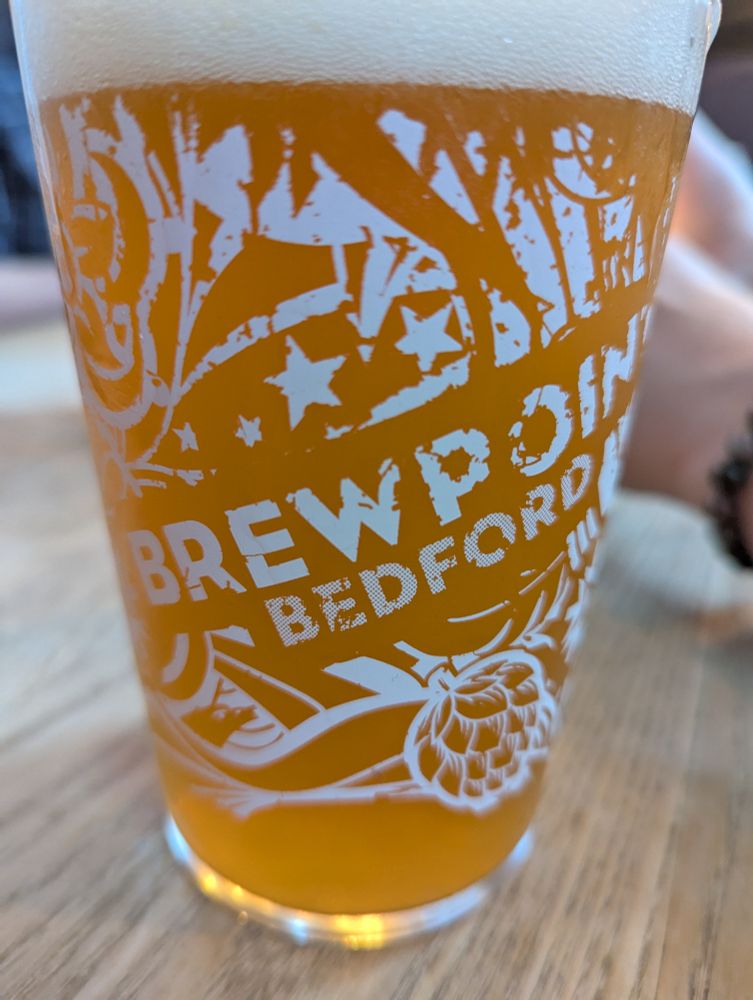A close up photo of a pint glass that's from a brewer called brewpoint Bedford
The pint is an amber orange colour on a wooden table with plenty of the wood grain showing 