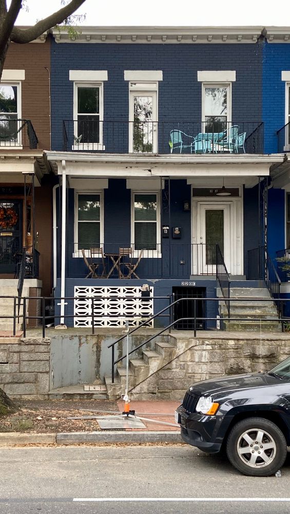 A rowhouse painted blue. A dark SUV is parked on the street in front. There is a porch above the front door with a light green table and chairs.