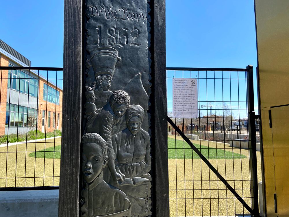 A post with Black figures depicted and 1862 above their heads. A grassy school yard is visible in the rear.