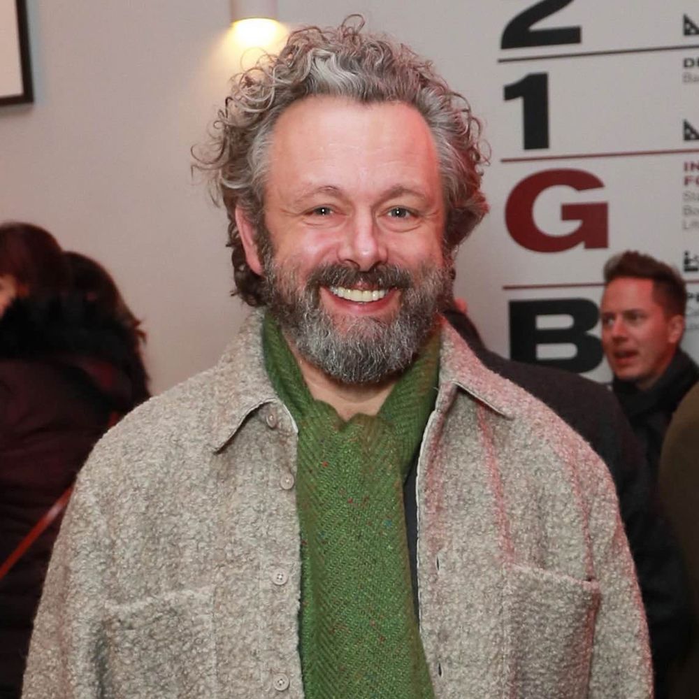 Michael Sheen, beaming at the camera. He's wearing a grey jacket/coat and a green scarf. 