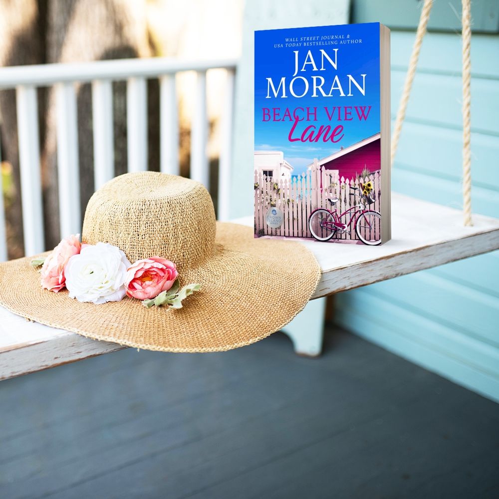 Image of Beach View Lane book by Jan Moran. Image is of the book and pretty sunhat on a porch swing. The book image is of a pink beach house and bicycle with a picket fence and the ocean beyond. 