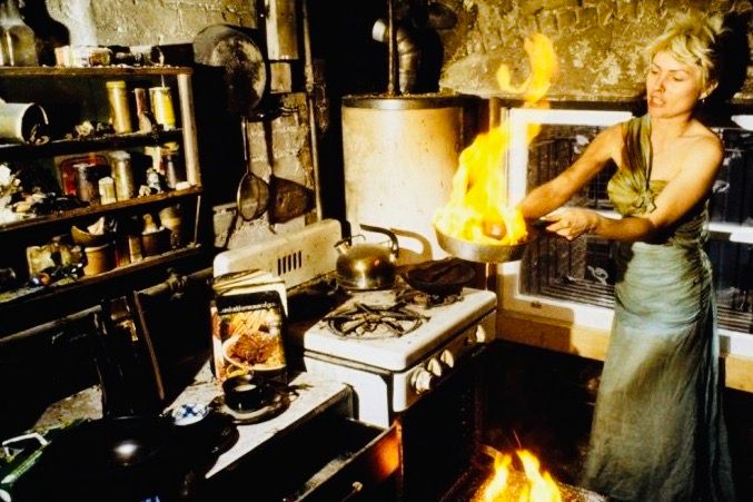 A dishevelled Debbie Harry in a long dress, in a dingy, fire-scarred kitchen, furnished with makeshift shelves and aged appliances, holding a burning frying pan out in front of her.