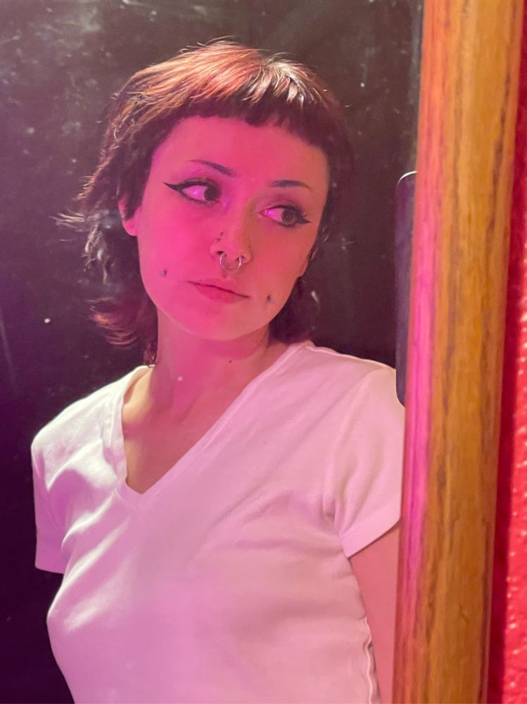 maple taking a mirror selfie in pink lighting, wearing a white shirt which is very rare. they are looking to the side at their phone camera, which is out of view. maple has short dark hair with mini bangs, tiny lil eyebrows, and face piercings (paired high nostrils & nostrils, septum, cheeks) 