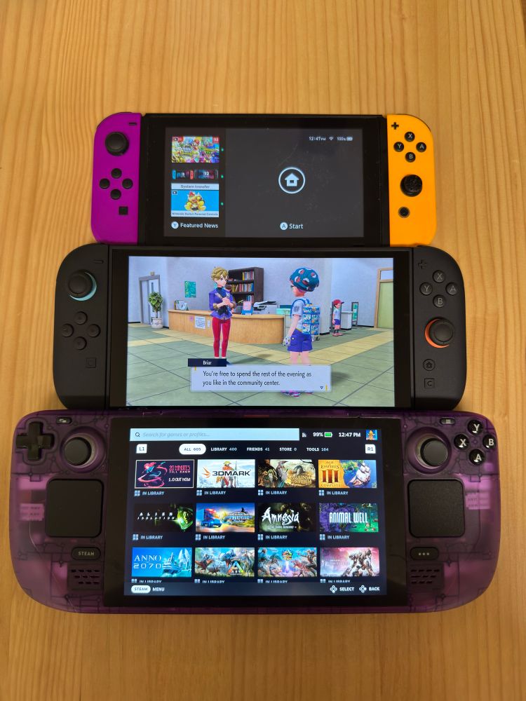 A Nintendo Switch one with purple and orange joycons, a new Nintendo Switch 2 playing Pokémon Violet, and a translucent purple Steam Deck stacked next to each other on a wooden table. The Steam Deck is the largest device, following by the Switch 2, and then the original Switch.