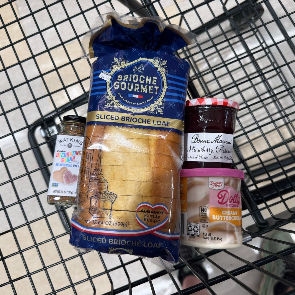 A bottle of decorating sugar, a loaf of slice brioche bread, a jar of strawberry preserves, and a can of buttercream icing in a shopping cart 