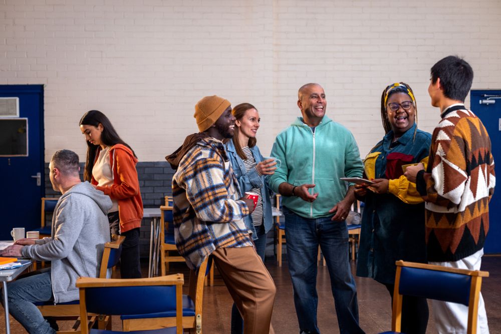 A diverse group of people are standing and chatting in a bright room with wooden chairs. One man in a plaid jacket holds a red cup, while another in a green hoodie laughs. A woman with glasses holds a tablet, engaging with a man in a geometric sweater. A woman and a man sit and work at a table nearby.