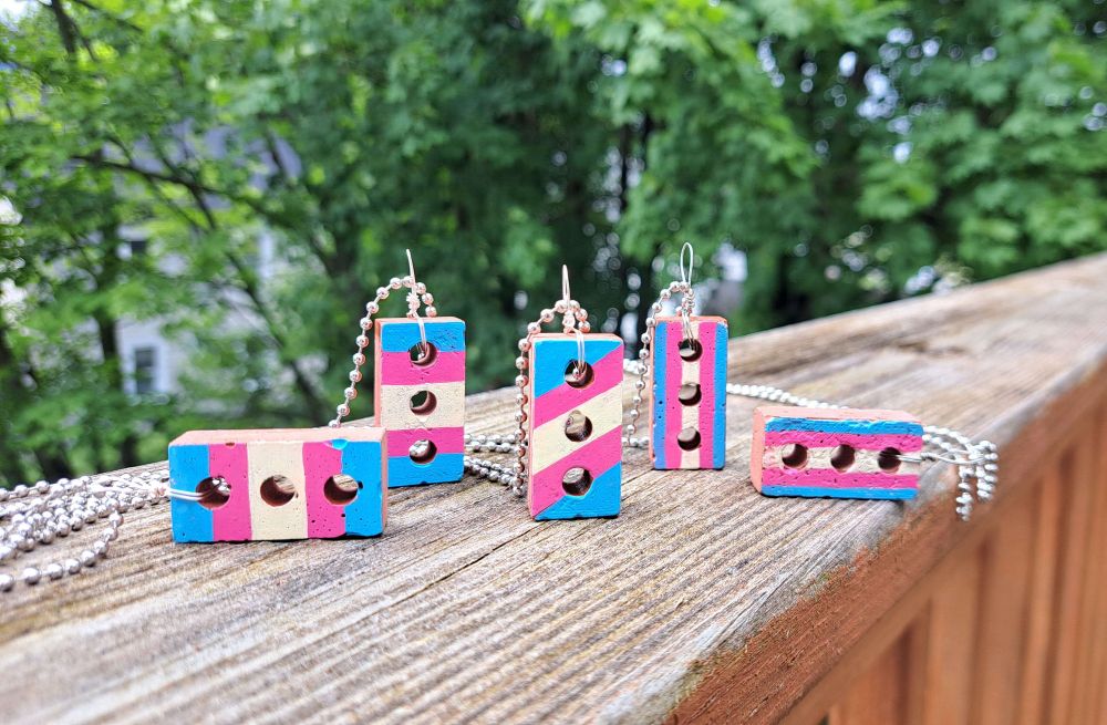 Clay brick necklaces hand-painted with the trans flag to represent the first brick thrown at Stonewall and in protest of transgender representation being removed from the Stonewall government page. The flag is portrayed horizontally, vertically, and diagonally on different bricks and each is on a metal chain.