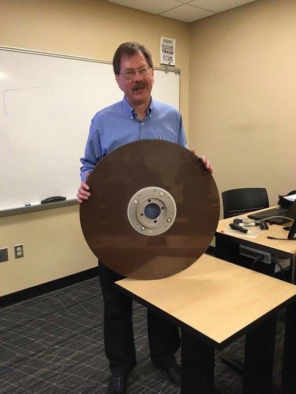 A man in a blue shirt holds up a very large, vintage hard disk platter that is 10MB in size in a office setting.
