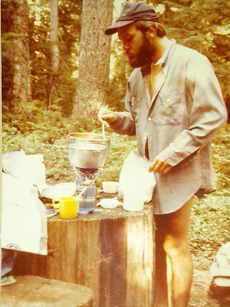 Me when my beard was still dark wearing a light long sleeve shirt and shorts. Cooking dinner on a couple single burner camp stove using a stump for a table