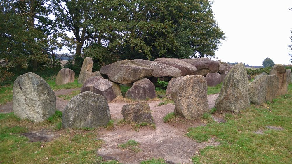 Hunebed D50, a neolithic burial structure in a grassy field surrounded by trees, near the Dutch town of Noord-Sleen. An inner ring of half buried large rocks is covered by horizontal flat rocks. This structure is surrounded by a second ring of half-buried rocks. Initially these rocks would have been covered by a mound of soil. Many hunebedden were built east-west with the entrance facing south.