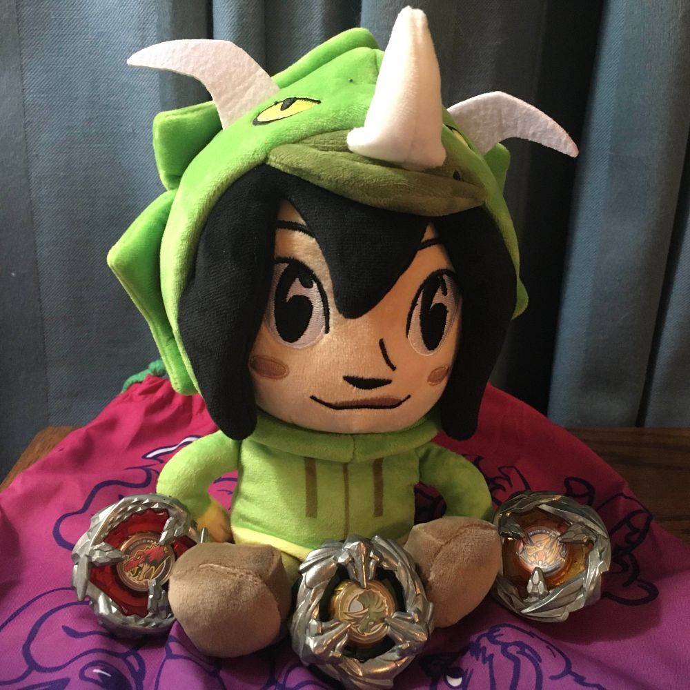 marketable plushie of Dina from Dumbing of Age, hanging out with some dinosaur-themed Beyblades (from left to right: TyrannoBeat, TriceraPress, and PteraSwing)
