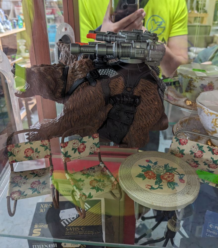 A bad ass war bear with lasers strapped to its back in front of a quaint mini doll table set.