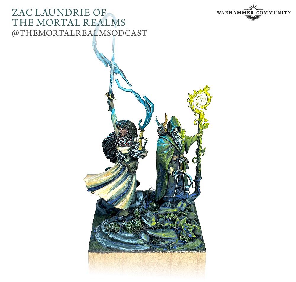 A miniature diorama. Two miniatures stand on alongside a small pond, in the grass.

On the left is a miniature of a woman in a light dress. In one hand she holds a jug and the other a sword. Water magically flows from the jug to meet her outstretched blade.

On the right is a robed mage in green. He holds a sickle on one hand and a staff of living vines in the other. On his shoulder sits an owl sprite.