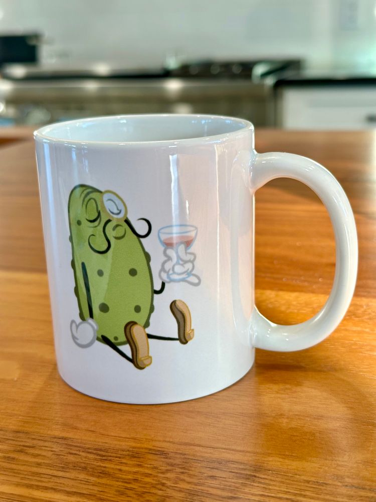 Shows a mug sitting on a walnut wood counter. The mug features an anthropomorphic Rather Dapper pickle with a monocle on the side drinking from a martini glass.