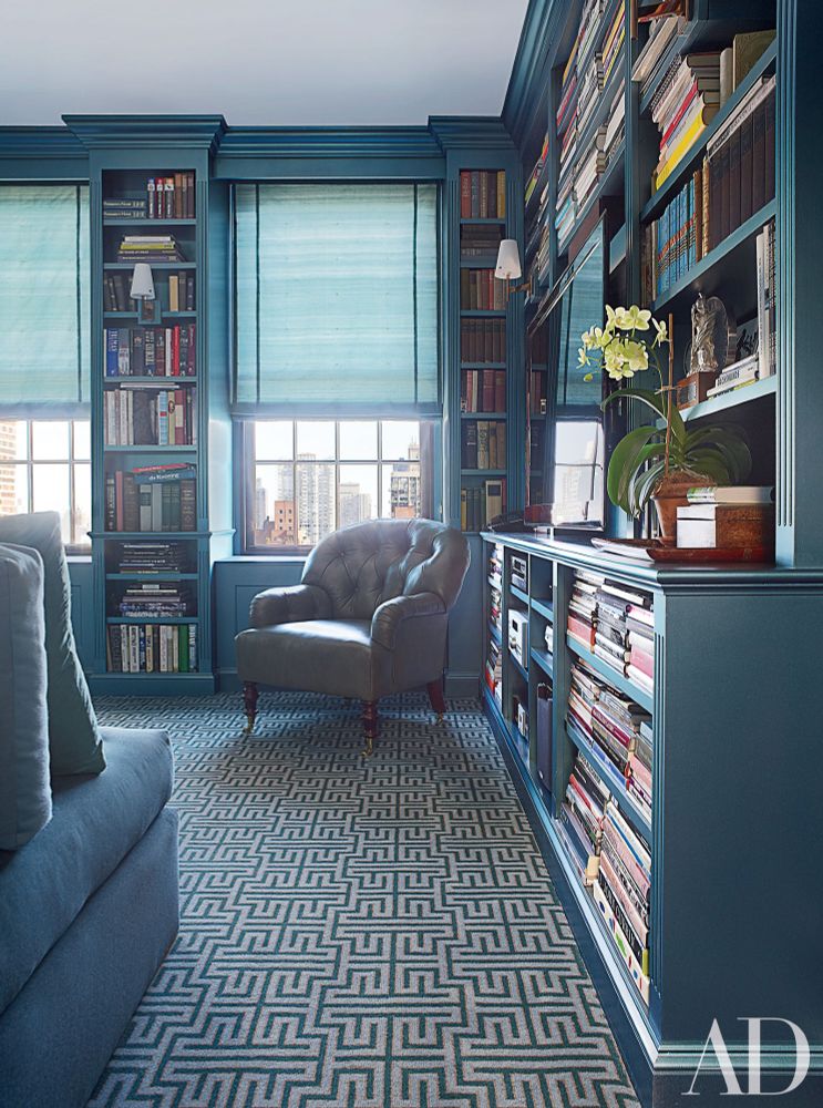 Manhattan private home library, walls lined with blue bookshelves, also between windows, matched by geometric carpet in blue and white, blue sofa and blinds, white armchair