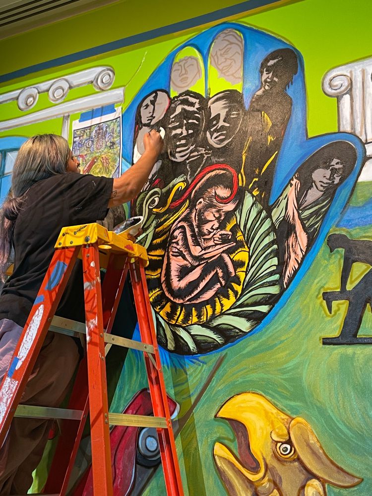 Bright green colored mural with an eagle and a black hand with kids faces being painted by a person with long hair on a ladder