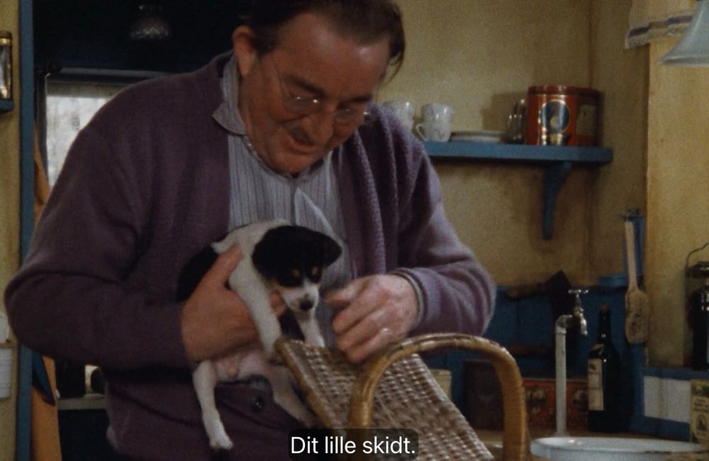 screenshot fra den danske serien ‘Matador’, hvor grisehandler Larsen tar opp en foxterrier fra en kurv med lokk. Han sier «Dit lille skidt.»