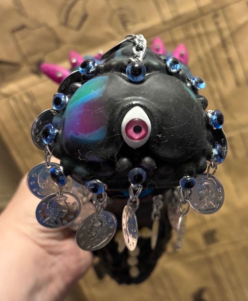 a central pink eye wmbwded in a ritual object.  decorated with dangling coins, teddy bear eyes, teeth and fingernails