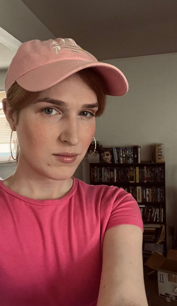 amanda is wearing a pink top and lulu pencil hat, with hoop earrings. behind her are the various films and tv shows she owns on home video.