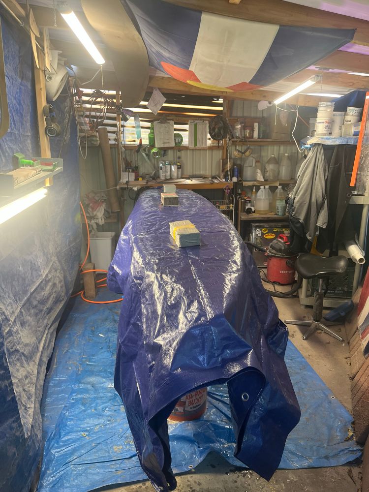 The PNerves surfboard shack (a small room with a tarp floor and plastic roof). The shack has several shelves and things hanging from the walls. A tarp covered surfboard sits on the racks. 