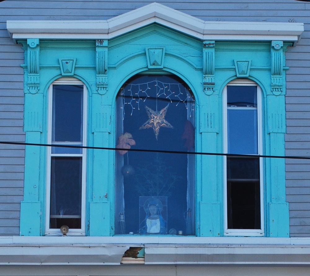 Photo of three windows on the second floor of a Victorian building. The paint on the elaborate trim is turquoise blue. There is a light in the shape of a star hanging in the center window, and a small stained glass depiction of Mary. 