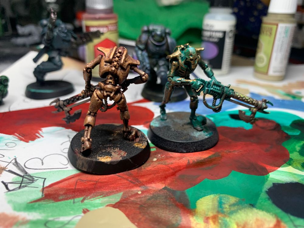 Two necrons: skeleton robots with huge guns. One is gold with red undertones and crackled paint. The other is brass with blue and green corrosion 