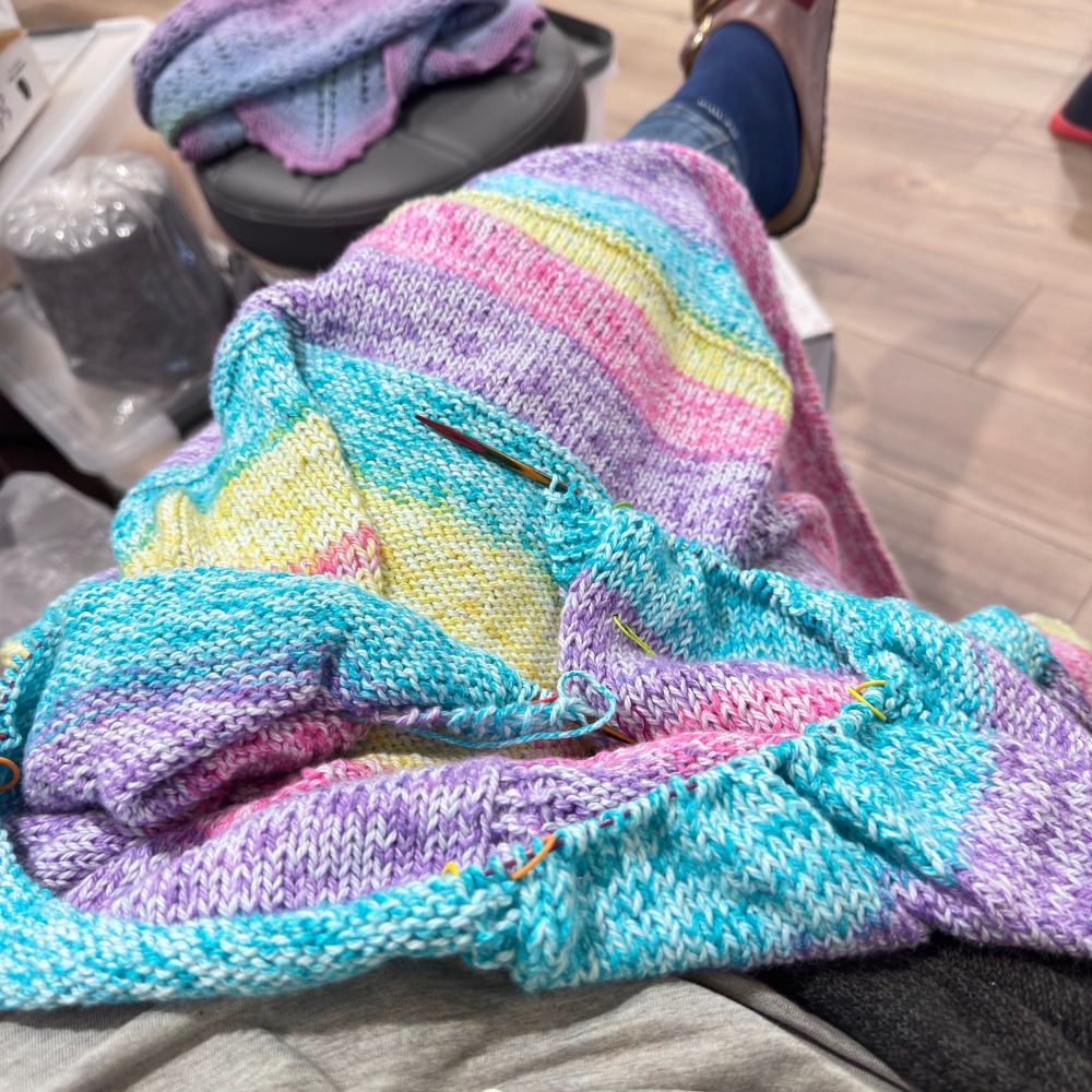 A small rainbow coloured blanket covers a woman’s lap, knitting needles on pause for a bit. 