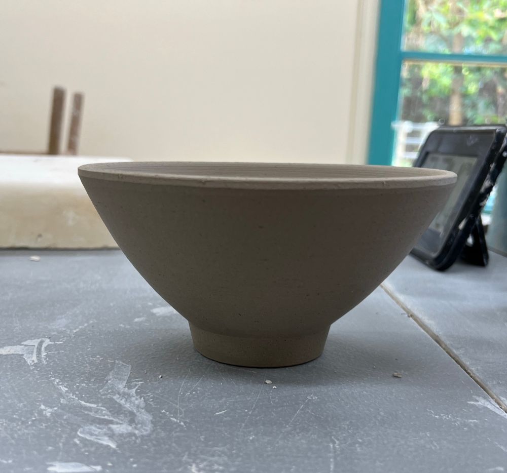 A greenware ceramic bowl that has just been trimmed, sitting on a grey table. It has a narrow foot and flares out wide. 
