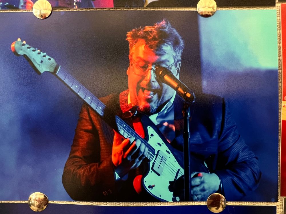A photo surrounded by thumbtacks attached to a cubicle wall. It depicts a guitarist (John Flansburgh of they might be giants) looking down at his guitar with his mouth open in seeming delight. 