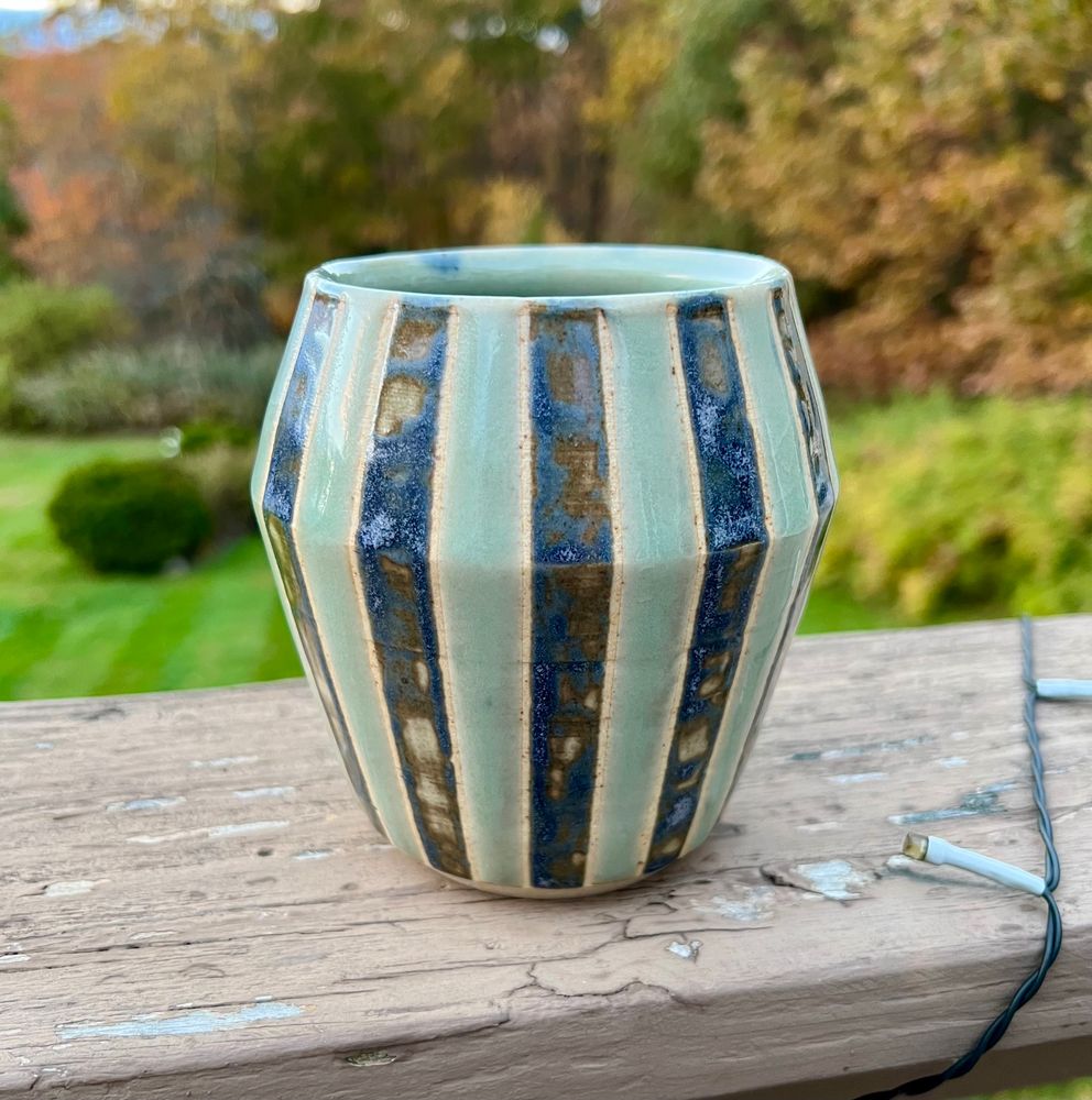 An angular ceramic cup sitting on a deck railing. It had alternating stripes of light and dark blue, with thin lines of bare clay on the borders of each colored line. The dark blue glaze is rather patchy. 