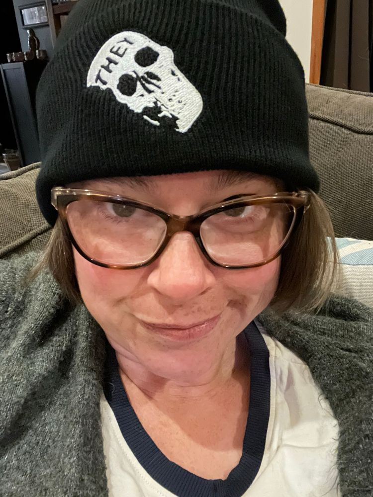 A woman in glasses wearing a black toque with a patch on it of a white skull with “THEY” written across it 