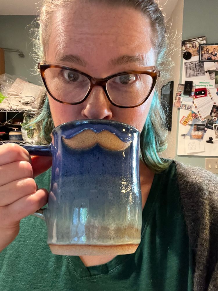 A woman in glasses holding a blue mug up to her mouth. The mug has a mustache on it and it makes her look like she has one. 