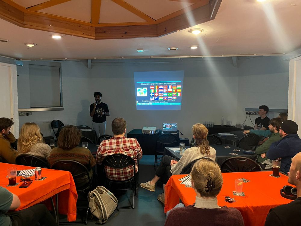 A pint of science volunteer introducing the evening. They are standing in front of a screen with a small crowd in front of him.
