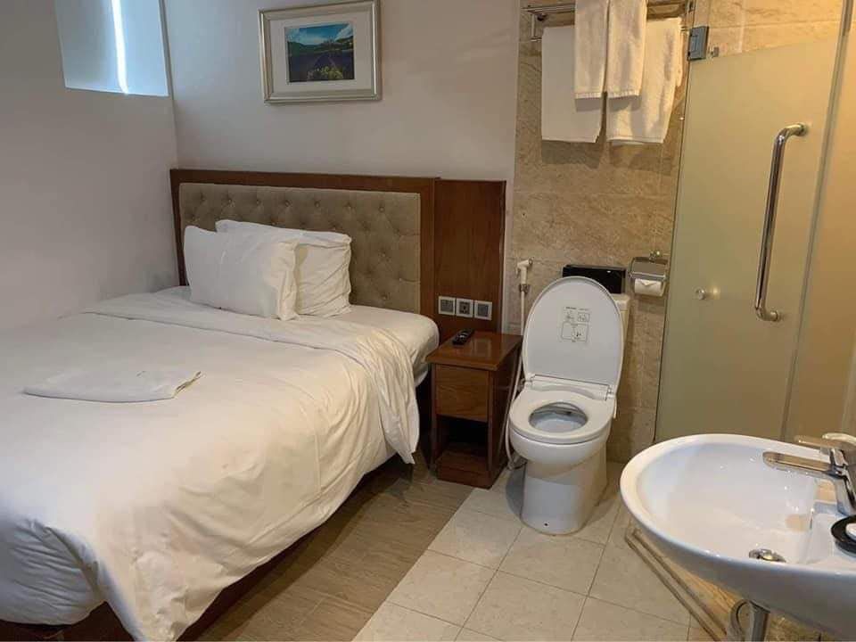 Das Bild zeigt ein modernes Schlafzimmer in neutralen Farben. In der Mitte des Raums steht ein Einzelbett mit weißer Bettwäsche. Direkt rechts vom Bett befindet sich eine Toilette ohne Trennwand, ganz dicht am Bettrand. Neben der Toilette stehen einige Hygieneartikel bereit. Der Raum ist hell beleuchtet und besitzt eine minimalistische Einrichtung: Am Fenster gegenüber steht ein kleiner Nachttisch. Es gibt keine weiteren Möbel oder Deko im Raum. Die Gesamtatmosphäre wirkt schlicht und ungewöhnlich, da die Toilette sehr nah und offen neben dem Bett platziert ist.
