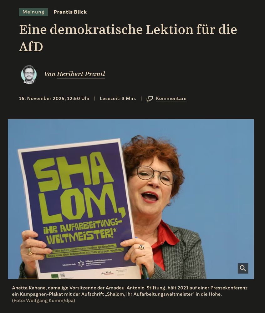 Auf dem Bild sieht man Anetta Kahane, die ehemalige Vorsitzende der Amadeu-Antonio-Stiftung. Sie hat kurze, lockige, rötlich-braune Haare und trägt eine Brille sowie ein kariertes graues Jacket über einer roten Bluse. Sie hält ein Plakat hoch, das in großen, grünlichen Buchstaben das Wort „SHALOM“ zeigt. Darunter steht in kleineren gelben Buchstaben: „Ihr Aufarbeitungsweltmeister!“. Das Plakat scheint von einer Kampagne gegen Antisemitismus zu stammen. Anettas Gesichtsausdruck ist lebhaft, sie scheint zu sprechen oder zu lachen. Der Hintergrund ist einfarbig blau. Unter dem Bild befindet sich eine Bildunterschrift, die erklärt, dass das Foto 2021 auf einer Pressekonferenz gemacht wurde und von Wolfgang Kumm/dpa ist.
