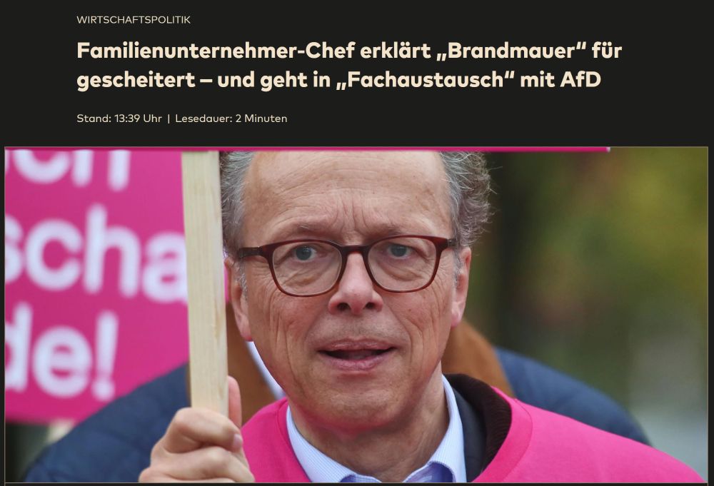 Auf dem Bild ist ein Mann mittleren Alters zu sehen, der eine rot-braune Brille trägt. Er hat graues, leicht gelocktes Haar und trägt einen pinken Pullover über einem weißen Hemd. Er hält ein Schild oder einen Holzstab in der Hand, dessen Aufschrift nicht sichtbar ist. Im Hintergrund erkennt man eine weitere Person und ein großes, pinkes Schild mit weißer Schrift, von der nur Ausschnitte zu lesen sind. Der Mann blickt frontal in die Kamera. Die Szene wirkt wie eine Aufnahme im Freien, vermutlich bei einer Demonstration oder Veranstaltung.