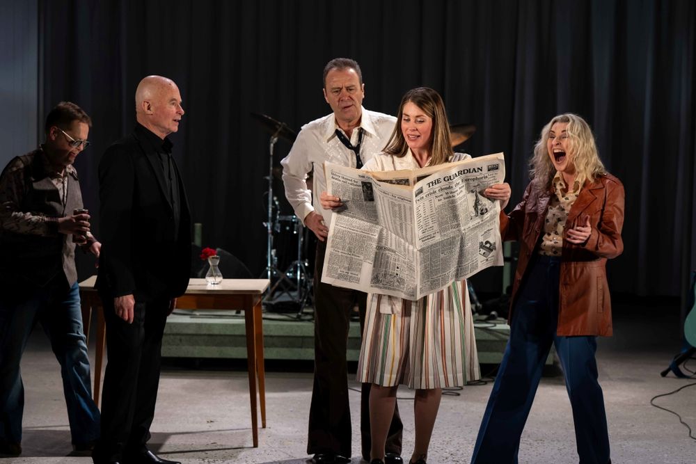 Georgina Fairbanks as Irene Knight reads a broadsheet of The Guardian as the band stand behind her smiling