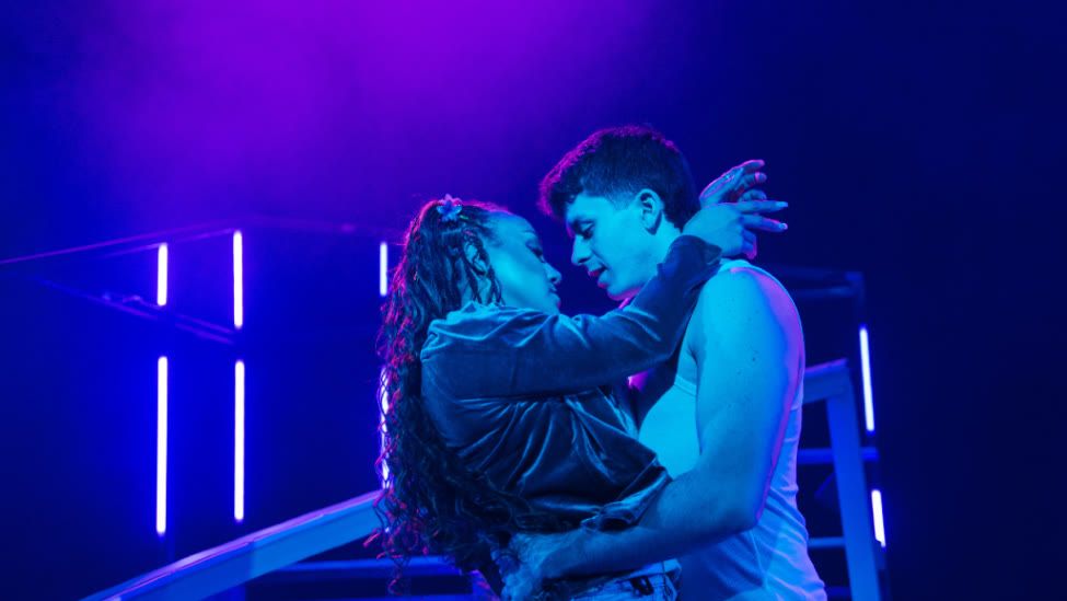 Earth (Danielle Steers) and Humanity (Tobias Turley) stand in a close embrace. Earth's arms rest of Humanity's shoulders and his wrap around her waist.