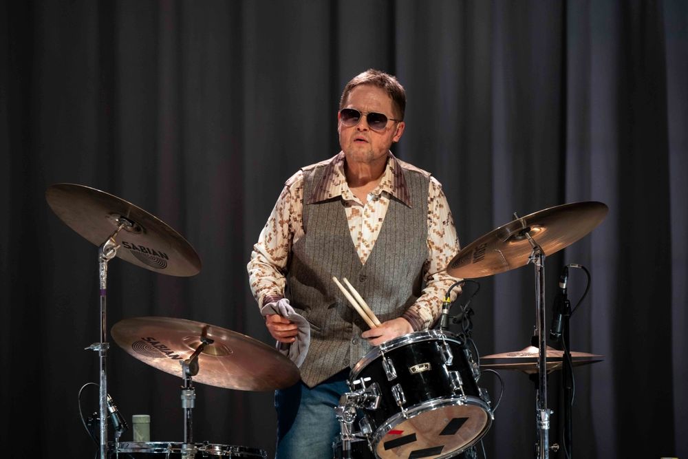 David John as Big Figure wears a waistcoat and sunglasses sat behind a drumkit